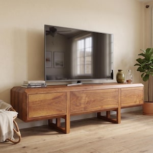 Puede incluir: Un televisor de pantalla plana grande se encuentra sobre una consola de medios baja de madera. La consola tiene un acabado de madera natural y c&aacute;lido con tres cajones. Una peque&ntilde;a planta en una maceta de terracota y botellas decorativas est&aacute;n a la derecha. Una silla de mimbre con una manta est&aacute; a la izquierda.