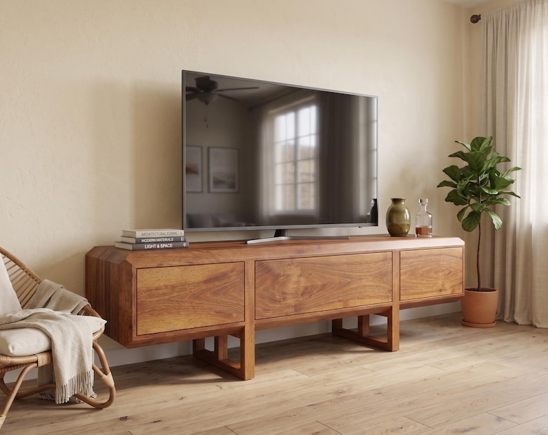 Puede incluir: Un televisor de pantalla plana grande se encuentra sobre una consola de medios baja de madera. La consola tiene un acabado de madera natural y c&aacute;lido con tres cajones. Una peque&ntilde;a planta en una maceta de terracota y botellas decorativas est&aacute;n a la derecha. Una silla de mimbre con una manta est&aacute; a la izquierda.