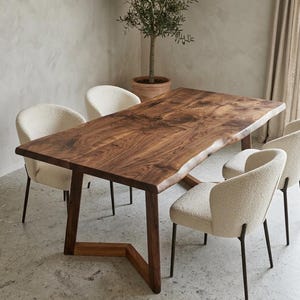 Op de afbeelding: Een rechthoekige eettafel met een donkerhouten blad met natuurlijke boomstamrand en een V-vormig houten onderstel, omringd door vier crèmekleurige stoelen van gestructureerde stof met dunne zwarte metalen poten in een eetkamer.
