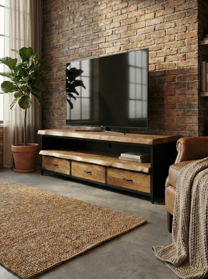 Puede incluir: Un mueble de televisi&oacute;n rectangular con una superficie y estante de madera natural de borde vivo, sostenido por un marco de metal negro. La unidad incluye tres cajones de madera con tiradores de metal oscuro para almacenamiento.
