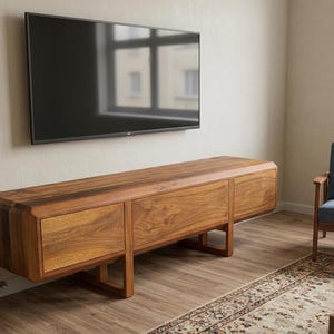 Puede incluir: Un mueble de TV moderno de madera con tres cajones y forma rectangular. El mueble est&aacute; hecho de madera de tonos c&aacute;lidos y est&aacute; sostenido por patas anguladas. Un televisor de pantalla plana negro grande est&aacute; montado encima.