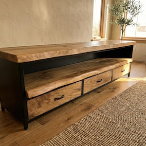 Puede incluir: Un mueble multimedia bajo con una superficie de madera natural de borde vivo y un estante abierto a juego. La base est&aacute; construida en metal negro e incluye tres cajones con tiradores negros delgados, ofreciendo espacio de almacenamiento.