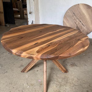 May include: A round wooden table with a natural wood grain finish. The table has a unique, organic edge and is supported by a central leg and three angled legs. The table's diameter is approximately 48 inches.