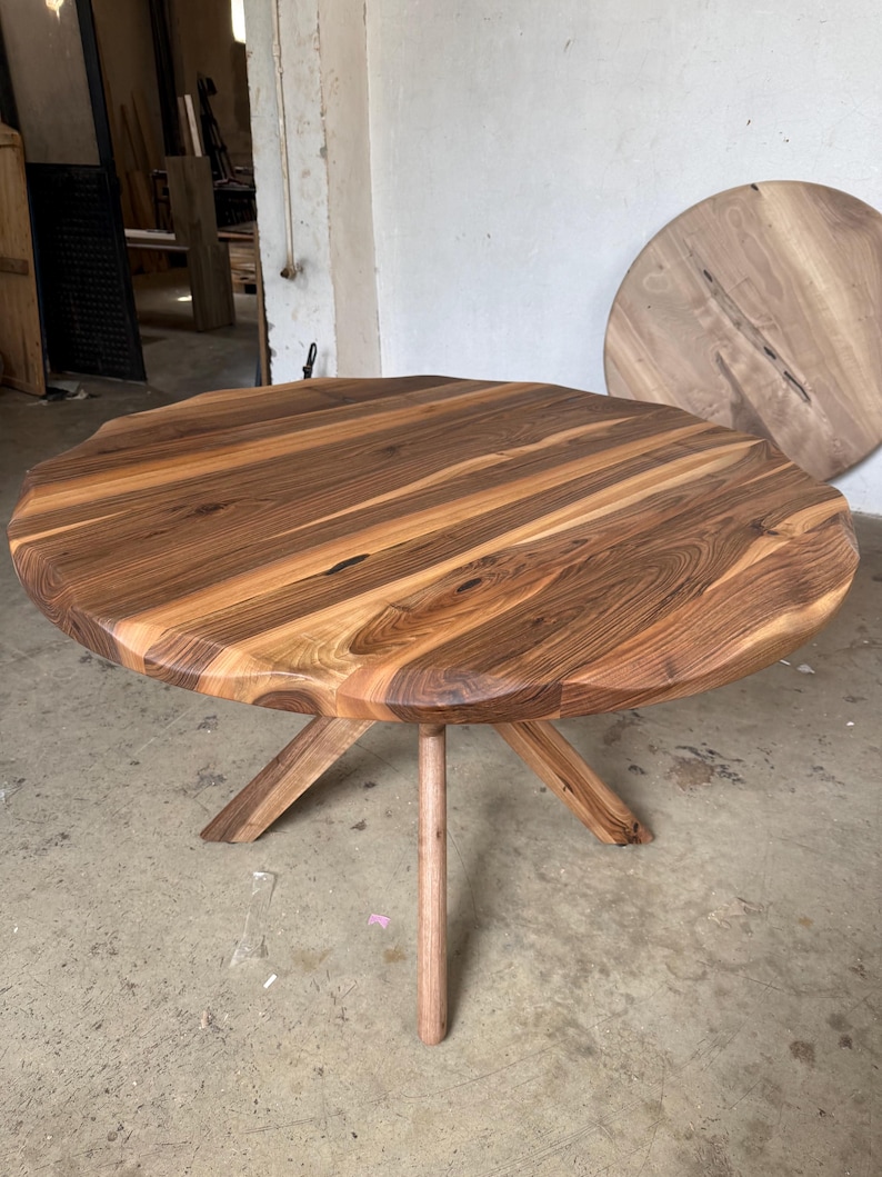 May include: A round wooden table with a natural wood grain finish. The table has a unique, organic edge and is supported by a central leg and three angled legs. The table's diameter is approximately 48 inches.