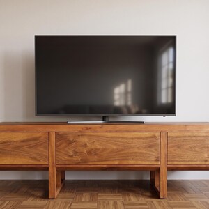 Puede incluir: Un mueble de TV moderno de madera con un televisor de pantalla plana grande encima. El mueble tiene un acabado de madera natural y c&aacute;lido con tres cajones y un dise&ntilde;o minimalista. El televisor es negro y refleja la luz de la habitaci&oacute;n.