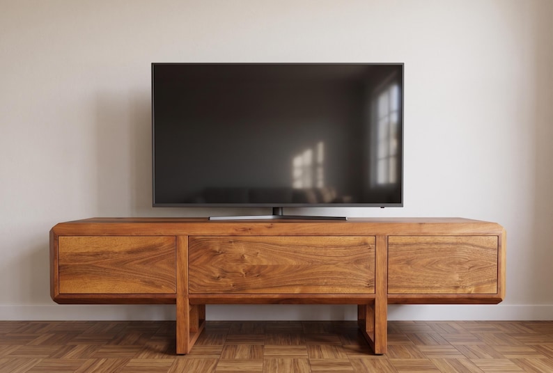 Puede incluir: Un mueble de TV moderno de madera con un televisor de pantalla plana grande encima. El mueble tiene un acabado de madera natural y c&aacute;lido con tres cajones y un dise&ntilde;o minimalista. El televisor es negro y refleja la luz de la habitaci&oacute;n.