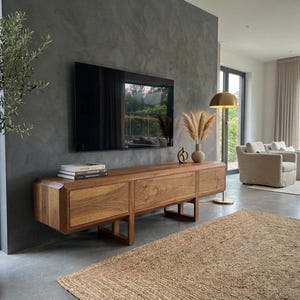 Puede incluir: Sal&oacute;n moderno con un televisor grande y oscuro montado sobre una consola de medios de madera. La consola tiene tres cajones y un dise&ntilde;o minimalista. Una alfombra decorativa y una planta en maceta a&ntilde;aden calidez al espacio. La consola mide unos 180 cm.