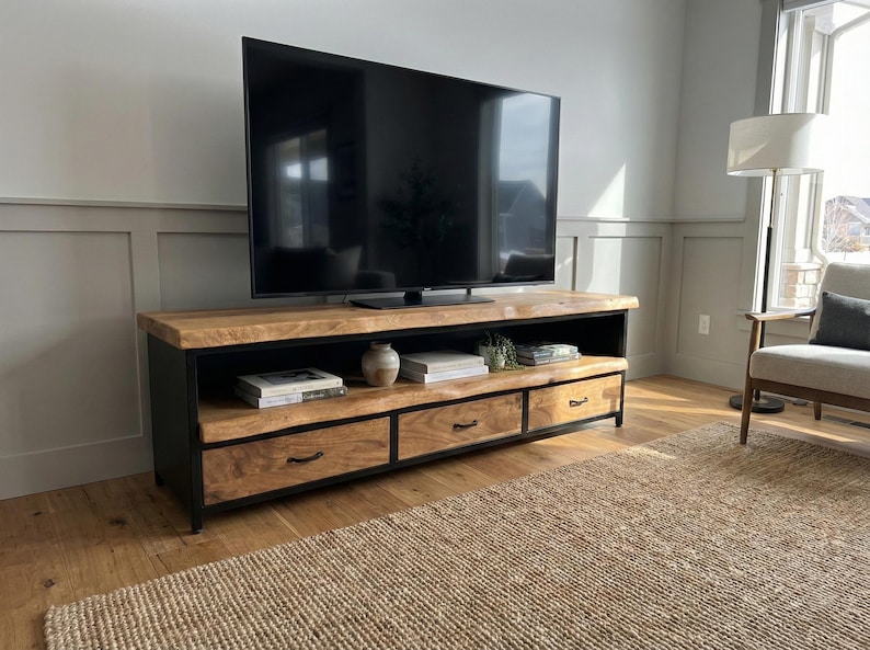 Puede incluir: Un mueble de televisi&oacute;n rectangular con una superficie y estante de madera natural, sostenido por un marco de metal negro. La unidad incluye tres cajones con tiradores de metal negro, proporcionando espacio de almacenamiento bajo el estante abierto.