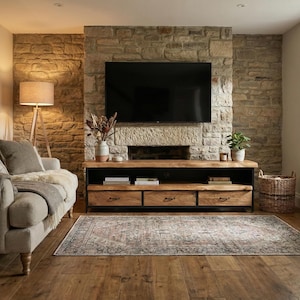 Puede incluir: Un mueble de televisi&oacute;n rectangular de madera con una superficie de borde natural, tres cajones con tiradores de metal oscuro y un estante abierto. La unidad cuenta con un marco de metal negro y se sit&uacute;a contra una pared de piedra bajo un televisor.
