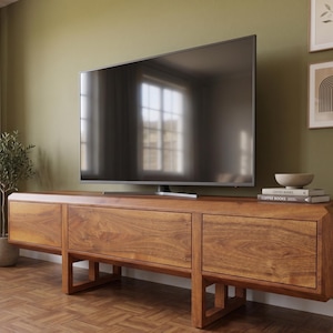 Puede incluir: Un televisor grande con pantalla oscura se encuentra sobre una consola de medios baja de madera. La consola es de madera de tonos c&aacute;lidos con veta natural, con tres cajones. Se ve una peque&ntilde;a planta en maceta y obras de arte enmarcadas.