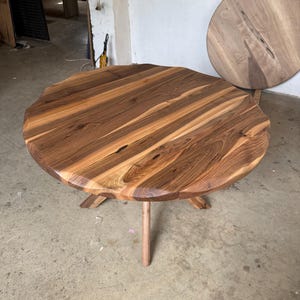 May include: A handcrafted, round wooden table with a natural edge. The tabletop is made of multiple wood planks, showcasing a mix of light and dark brown tones. The table stands on three angled legs and a central support.
