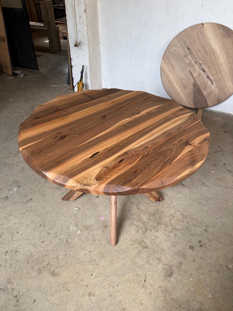 May include: A handcrafted, round wooden table with a natural edge. The tabletop is made of multiple wood planks, showcasing a mix of light and dark brown tones. The table stands on three angled legs and a central support.