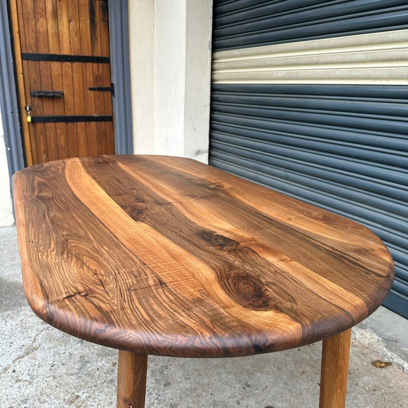 Pu&ograve; includere: Primo piano di un tavolo ovale in legno con finitura in legno naturale. Il tavolo ha una venatura unica e poggia su quattro gambe.