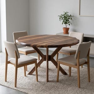 May include: A round wooden dining table with a dark brown finish, surrounded by four chairs with beige upholstered seats and light wood frames. The table sits on a beige rug, with a small potted plant in the background.