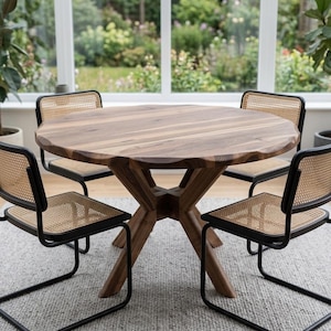 May include: A round wooden dining table with a unique, textured edge, supported by a cross-shaped base. Four chairs with black metal frames and woven cane backs surround the table. The room has a neutral color palette with a light gray rug.