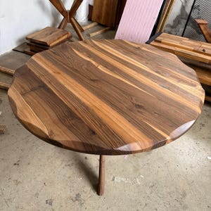 May include: A round wooden table with a unique, wavy edge. The tabletop is a mix of light and dark brown wood grains. It stands on a single, slender wooden leg. The table's diameter is approximately 40 inches.