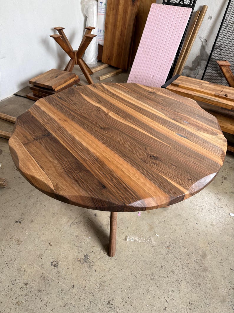 May include: A round wooden table with a unique, wavy edge. The tabletop is a mix of light and dark brown wood grains. It stands on a single, slender wooden leg. The table's diameter is approximately 40 inches.