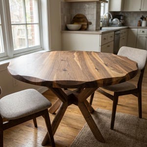 May include: A round wooden dining table with a unique, wavy edge. The table is supported by a crossed-leg base and is surrounded by two chairs with light-colored upholstered seats. The table sits on a woven rug in a well-lit kitchen.
