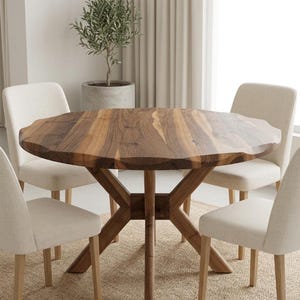 May include: A solid wood dining table with a unique, organic edge design. The table is supported by a sturdy, crossed-leg base. Four cream-colored upholstered chairs surround the table. A potted olive tree is in the background.