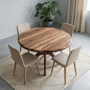 May include: A round wooden dining table with a natural wood grain finish, surrounded by four beige upholstered chairs. The table sits on a patterned rug, with a potted plant in the background. The room has neutral-toned walls and curtains.