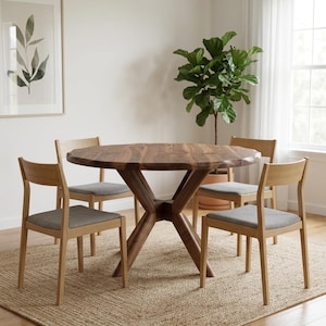 May include: A round wooden dining table with a dark finish, supported by a unique cross-shaped base. Four wooden chairs with gray upholstered seats surround the table. The scene is set on a woven jute rug.