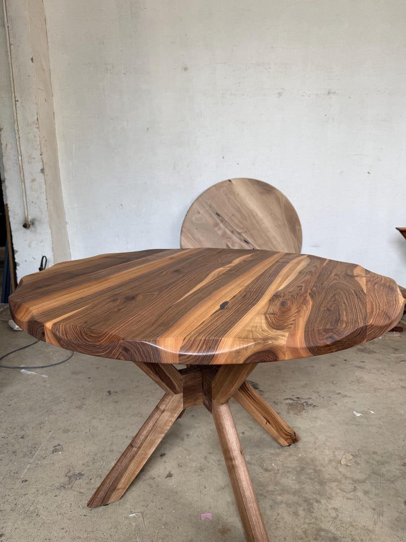 May include: A round wooden table with a natural edge and a starburst pattern on the tabletop. The table is supported by a wooden base with angled legs. The table is approximately 40 inches in diameter.