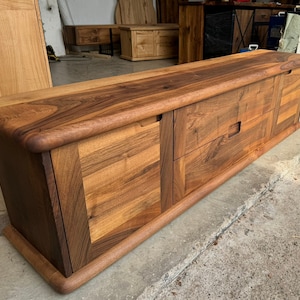 Può includere: Un mobile in legno artigianale con finitura marrone caldo. Presenta due cassetti e un'anta. Il mobile ha una finitura liscia e lucida, con bordi arrotondati. La venatura naturale del legno è visibile. Lunghezza di circa 180 cm.