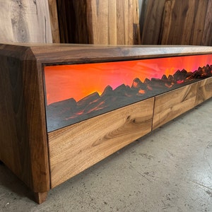 May include: A wooden media console with a striking sunset scene. The console features a dark wood top and sides, with a light wood front. The center panel displays a vibrant orange and pink sunset over a mountain range. The console has three drawers.