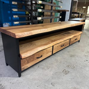 Puede incluir: Un mueble multimedia largo con una tapa gruesa de madera de color marr&oacute;n claro con borde natural y un estante abierto a juego. La base es de metal negro e incluye tres cajones con tiradores de metal negro, mostrando una veta de madera visible.