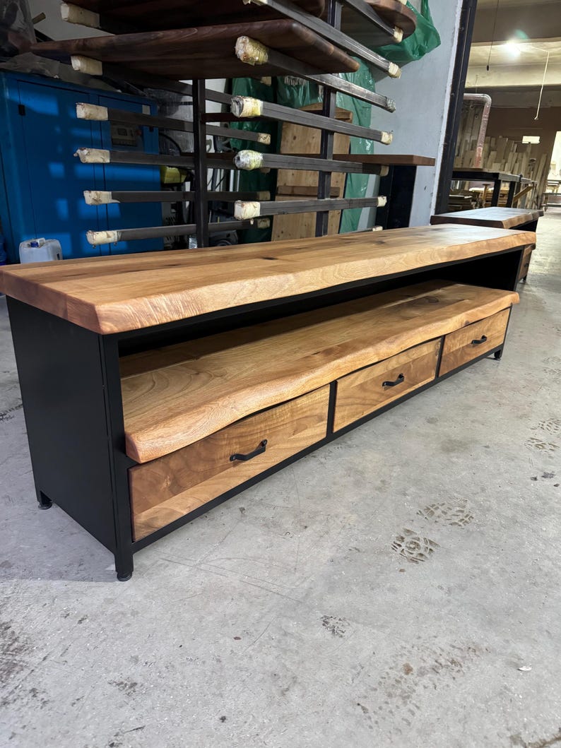 Puede incluir: Un mueble multimedia largo con una tapa gruesa de madera de color marr&oacute;n claro con borde natural y un estante abierto a juego. La base es de metal negro e incluye tres cajones con tiradores de metal negro, mostrando una veta de madera visible.