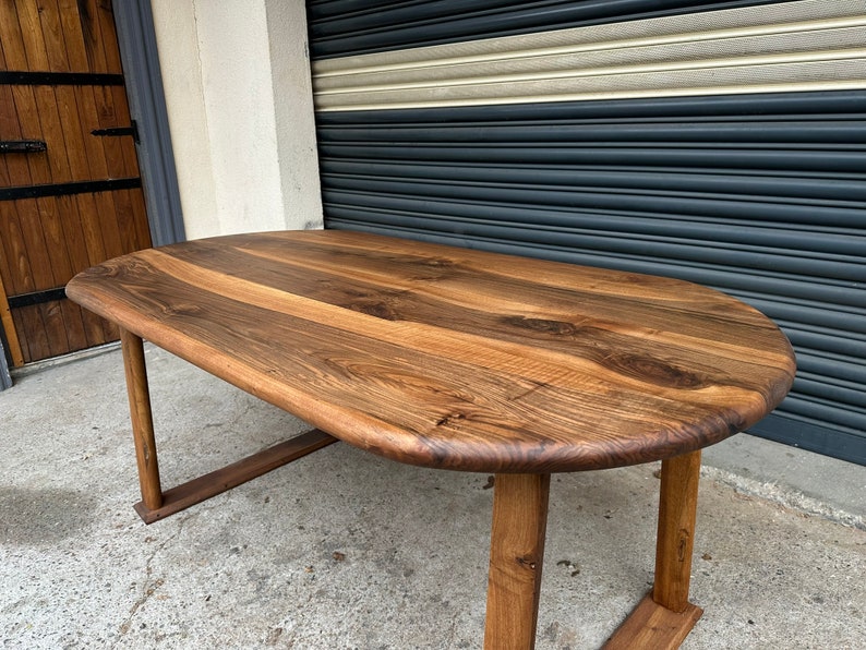 Pu&ograve; includere: Un grande tavolo ovale in legno con finitura in legno naturale. Il tavolo ha un piano spesso e solido e gambe semplici e robuste.