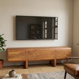 Puede incluir: Un sal&oacute;n moderno con un televisor de pantalla plana montado en la pared sobre un mueble de TV largo de madera clara con tres secciones. Una planta en maceta y un sill&oacute;n de cuero completan la escena.