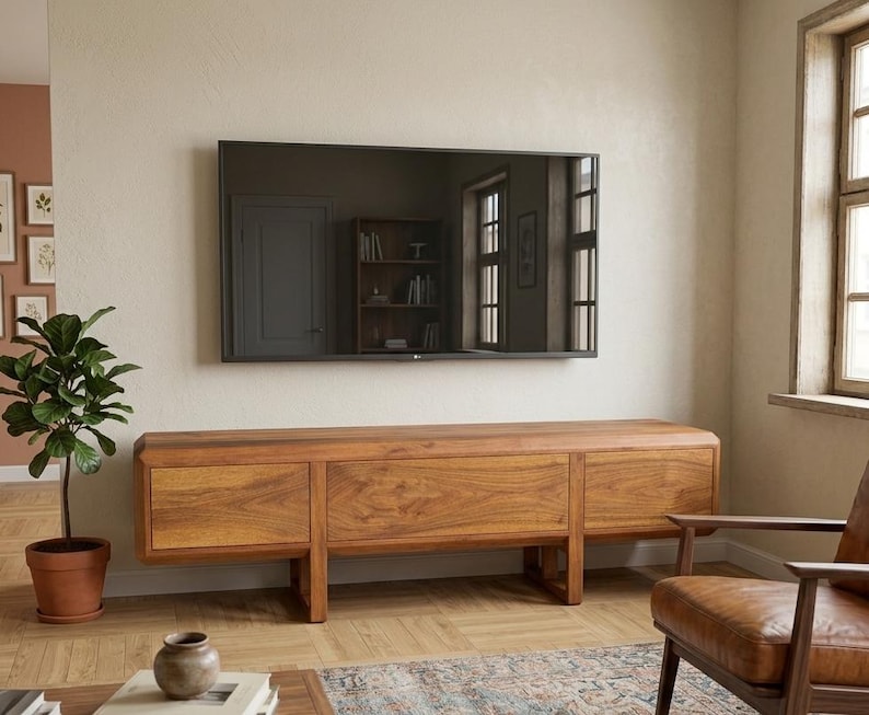 Puede incluir: Un sal&oacute;n moderno con un televisor de pantalla plana montado en la pared sobre un mueble de TV largo de madera clara con tres secciones. Una planta en maceta y un sill&oacute;n de cuero completan la escena.