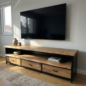 Puede incluir: Un mueble de televisi&oacute;n rectangular largo con una superficie de madera natural de borde vivo y tres cajones frontales con tiradores de metal negro. La unidad tiene un marco de metal negro, un estante de almacenamiento abierto y acabado veteado.