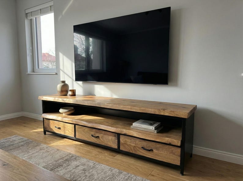Puede incluir: Un mueble de televisi&oacute;n rectangular largo con una superficie de madera natural de borde vivo y tres cajones frontales con tiradores de metal negro. La unidad tiene un marco de metal negro, un estante de almacenamiento abierto y acabado veteado.