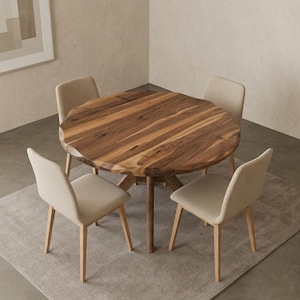 May include: A wooden dining table with a natural wood grain finish, surrounded by four beige upholstered chairs. The table sits on a light gray rug. A potted plant is in the background. The room has a neutral color palette.