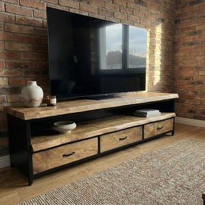 Puede incluir: Un mueble de televisi&oacute;n rectangular con una superficie de madera natural de borde vivo y dos estantes abiertos, sostenido por un marco de metal negro. La unidad incluye tres cajones inferiores con tiradores de metal negro para almacenamiento.