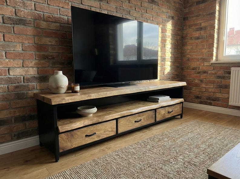 Puede incluir: Un mueble de televisi&oacute;n rectangular con una superficie de madera natural de borde vivo y dos estantes abiertos, sostenido por un marco de metal negro. La unidad incluye tres cajones inferiores con tiradores de metal negro para almacenamiento.