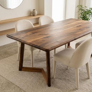 May include: A rectangular dining table with a dark brown wood grain surface and rounded corners, supported by a V-shaped wooden base. Three upholstered chairs with cream-coloured fabric and rounded backrests are positioned around the table.