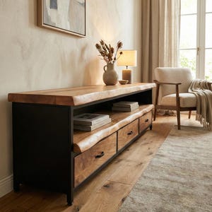 Puede incluir: Un mueble multimedia largo con estructura de metal negro y tres cajones con frentes de madera natural de borde vivo. La unidad incluye un estante abierto para almacenamiento y una superficie superior gruesa de madera a juego.