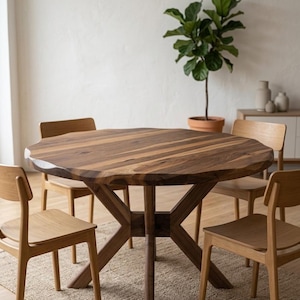 May include: A round wooden dining table with a natural wood grain finish, surrounded by four wooden chairs. The table has a unique, organic edge. A potted plant and neutral-toned decor are in the background.