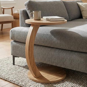 May include: A light wood, C-shaped side table with a round top and base. A mug and a book rest on the tabletop. The table is next to a grey sofa on a textured rug. The table's height is approximately 61 cm.