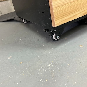 May include: A black and wood cabinet with four black wheels. The cabinet is on a gray concrete floor.
