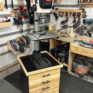 May include: A black drill press with a white head and a black metal base. The drill press is mounted on a wooden stand with four drawers. The stand has black metal drawer pulls.