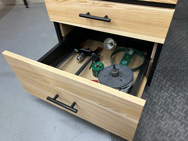 May include: Open drawer of a wooden cabinet with black hardware. The drawer is filled with various tools, including a drill bit, a hole saw, and a clamp.