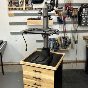 May include: A black and silver drill press with a black base and a wooden cabinet with four drawers. The drill press is mounted on the cabinet and has a black metal work surface. The cabinet has black drawer pulls. The drill press is in a garage setting with tools and shelves on the wall behind it.