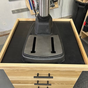 May include: A black drill press with a wooden base and three drawers. The drill press is mounted on a black platform with a wooden frame.