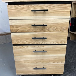 May include: A wooden cabinet with five drawers and black metal handles. The cabinet is on wheels and has a black top.