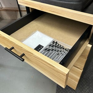 May include: A wooden drawer with a black handle is open, revealing a set of drill bits in a clear plastic container. The drawer is made of light wood with a black interior.