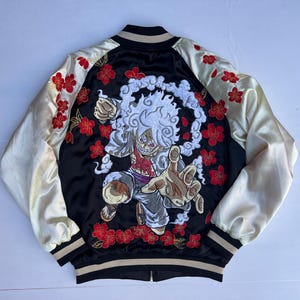 Può includere: Un giubbotto bomber in raso nero e crema con un grande ricamo dettagliato di un personaggio dei cartoni animati con capelli bianchi circondato da nuvole sul retro. Le maniche e il pannello posteriore sono decorati con fiori di ciliegio rossi.